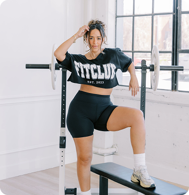 Woman in a gym by a bench press about to do a workout by The FITclub, an online community of real women and real bodies. 