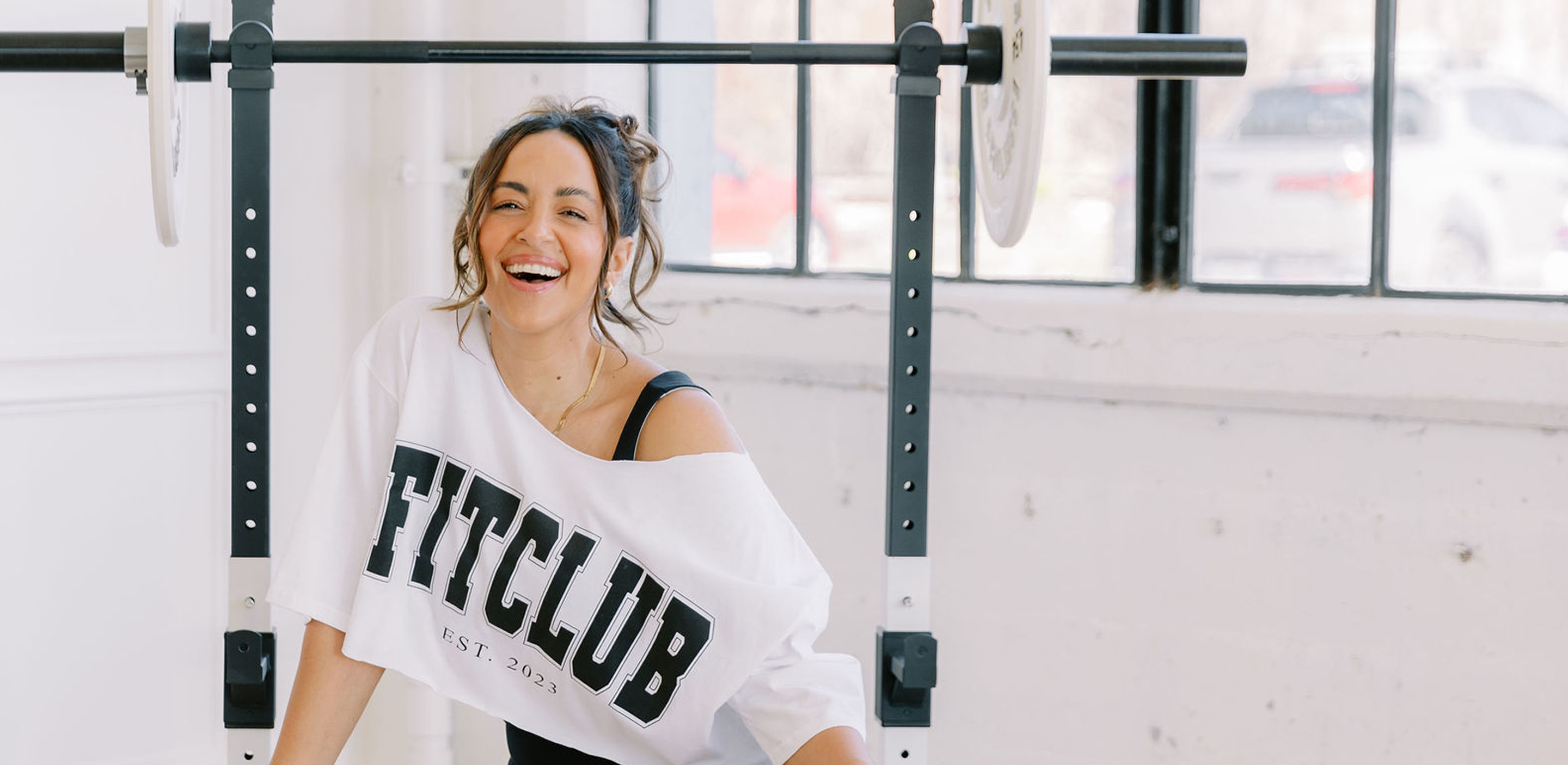 Woman sitting in a gym smiling next to weights wearing 'The FITclub' merchandise. 